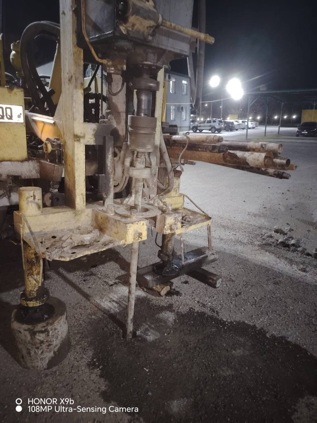 Yellow truck-mounted rotary drilling rig operating at night under street lamps near administrative buildings in Ambrolauri, Georgia