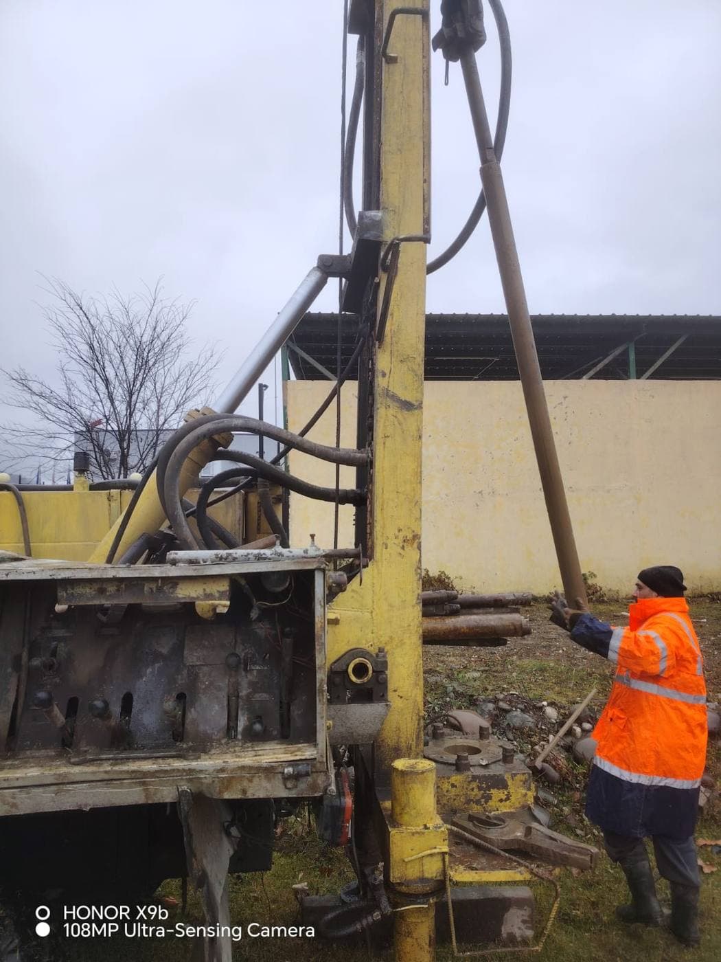 GGG technician in orange high-visibility jacket guiding drill rods on a rotary rig at the Ambrolauri administrative building site