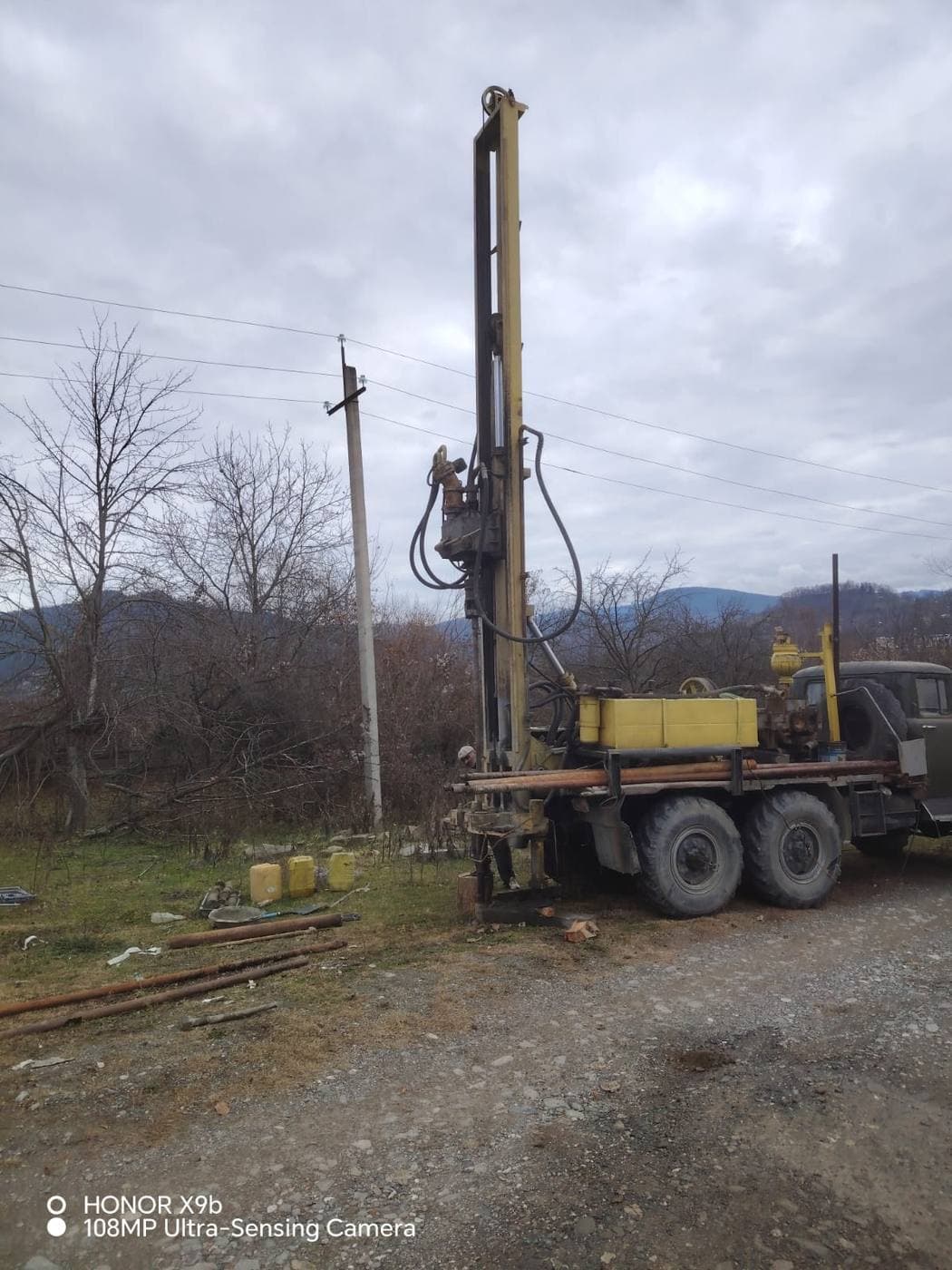 Older Soviet truck-mounted drilling rig at a rural site near Ambrolauri, Georgia, with utility pole and forested Caucasus hills behind